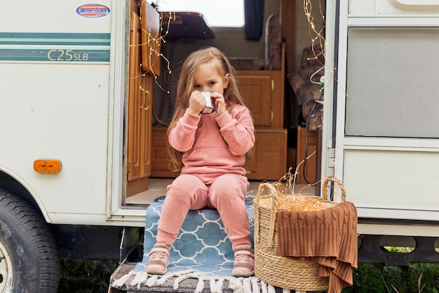 Schattig klein meisje zit op de trappen van een reisaanhanger en drinkt warme chocolademelk met marshmallows
