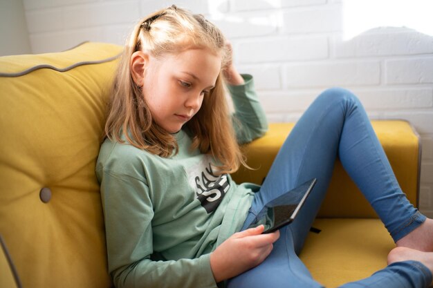 Schattig klein meisje zit op een bank en geniet van het spelen van een online game op een digitale tabletcomputer of luisteren naar een online webinar of het lezen van een boek