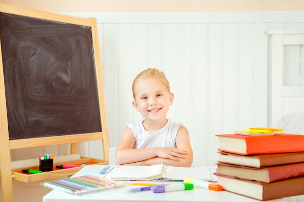 Schattig klein meisje, zittend door tafel met kleurpotloden en open voorbeeldenboek voor haar