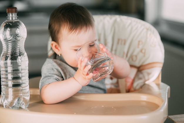 Schattig klein meisje, zittend in een kinderstoel in de keuken en drinkwater