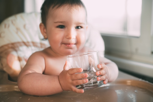 Schattig klein meisje, zittend in een kinderstoel in de keuken en drinkwater