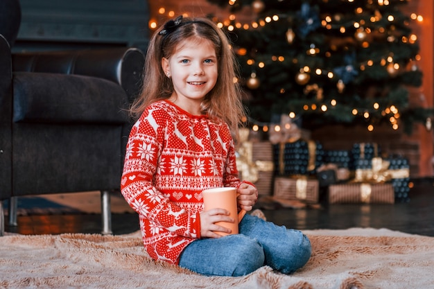 Schattig klein meisje zittend op de grond met een kopje drank in de kerst ingerichte kamer.