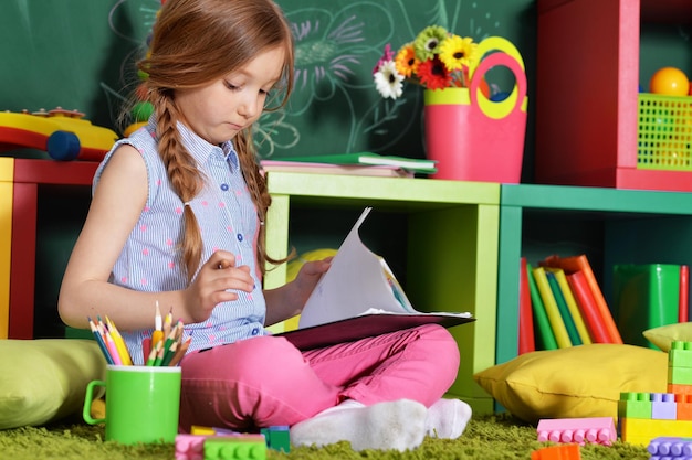 Schattig klein meisje zittend op de vloer en tekenen met potloden met schoolbord op achtergrond