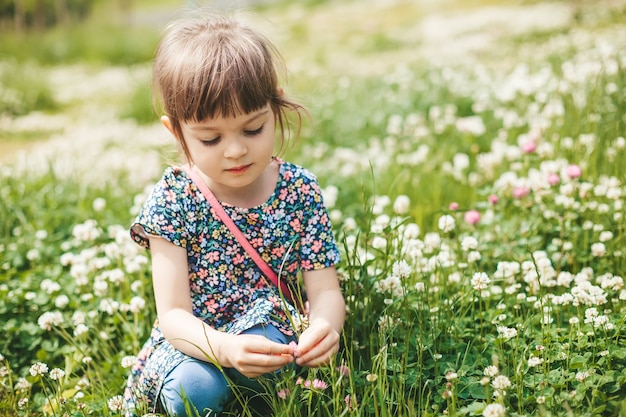 Schattig klein meisje zittend op een klaverveld, bloemen verzamelend en genietend van de zomer