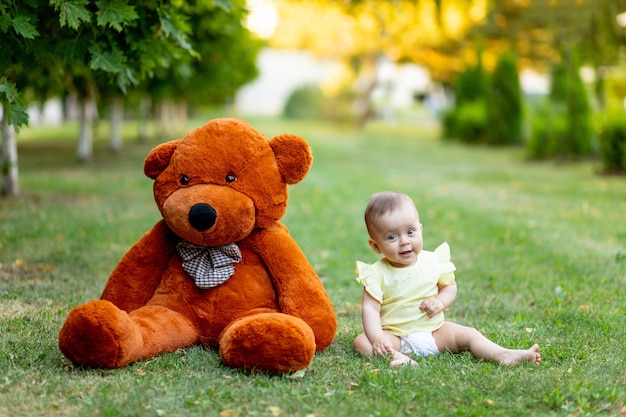 Schattig klein meisje zittend op groen gras met grote teddybeer in gele zomerjurk in de zomer.