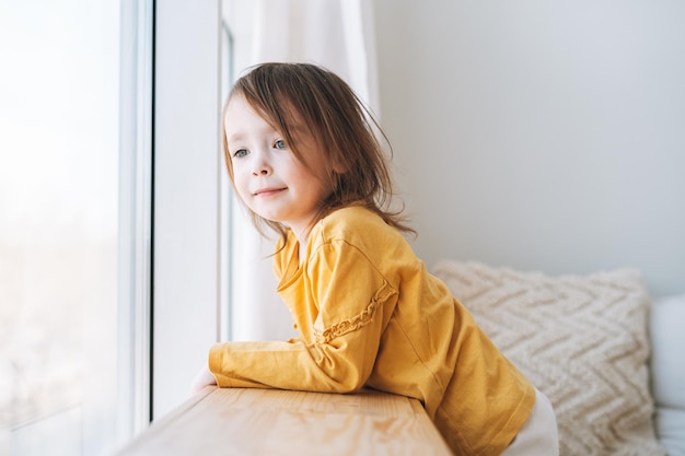 Schattig klein peutermeisje in gele longsleeve die thuis in het raam kijkt