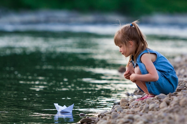 Schattig klein schattig blond meisje in blauwe jurk op rivieroever steentjes spelen met Witboek origami boot op blauw sprankelend bokeh water