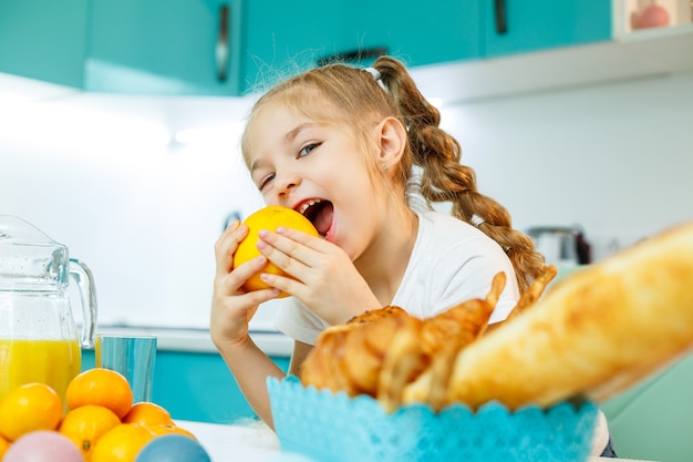 Schattig klein speels grappig meisje dat graag oranje sinaasappels vasthoudt, met keuken op de achtergrond