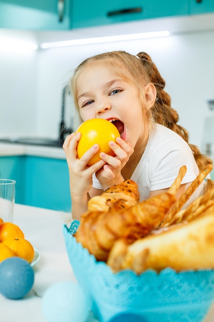 Schattig klein speels grappig meisje dat graag oranje sinaasappels vasthoudt, met keuken op de achtergrond