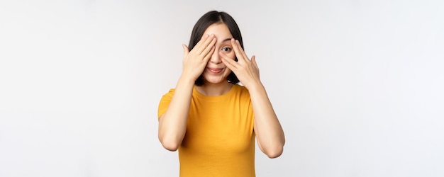 Schattig koreaans vrouwelijk model bedekt ogen met handen die door vingers gluren, nieuwsgierig kijken naar iets dat in gele t-shirt staat op witte achtergrond