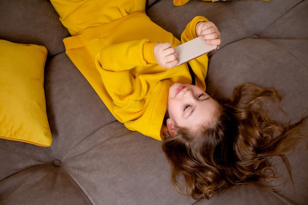 schattig lachend meisje in gele kleren ligt op haar rug op het bed en houdt een smartphone vast