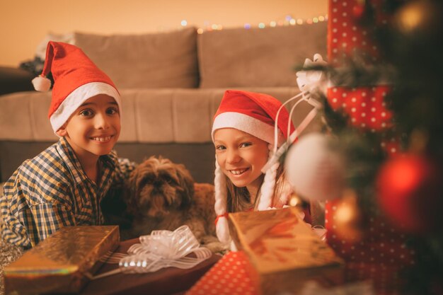 Schattig lachend meisje, jongen en hun kleine hond met veel cadeautjes thuis in een kersttijd. camera kijken.