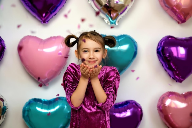 Schattig meisje blaast roze confetti in de vorm van harten uit haar handpalmen op een Valentijnsdagfeest