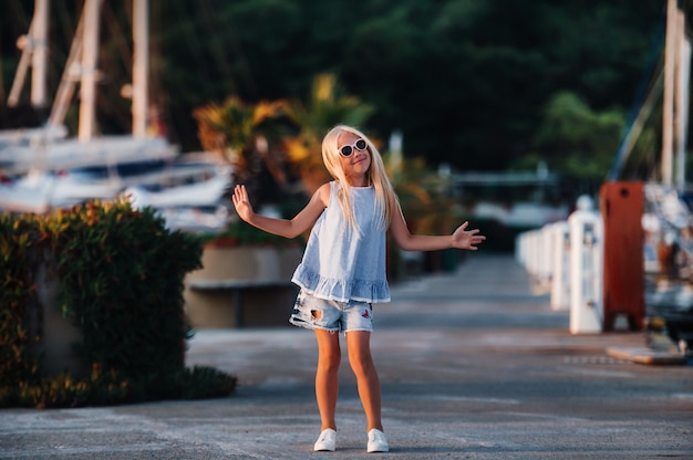 Schattig meisje in de buurt van de jachten in de zomer. Reizen, avontuur, boottochten met een kind voor een gezinsvakantie. Kinderkleding in de stijl van een zeeman, maritieme mode.