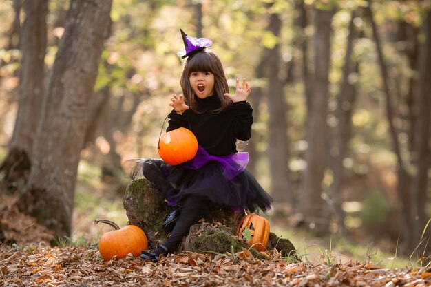 schattig meisje in heksenkostuum zit op boomstronk in herfstbos met pompoenlantaarn Jack Halloween