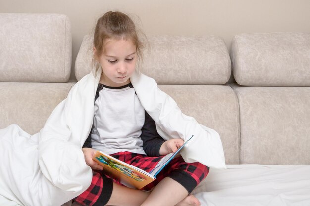 Schattig meisje in pyjama op het bed bedekt met een deken leest een boek