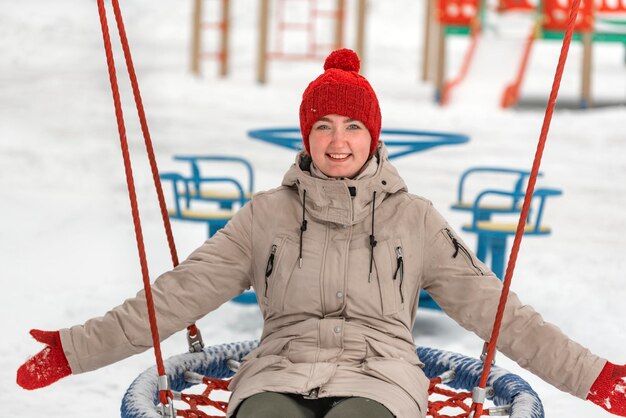 Schattig meisje in rode gebreide muts en wanten rijdt vrolijk op schommel op speelplaats Jonge vrouw heeft plezier in de winter buiten