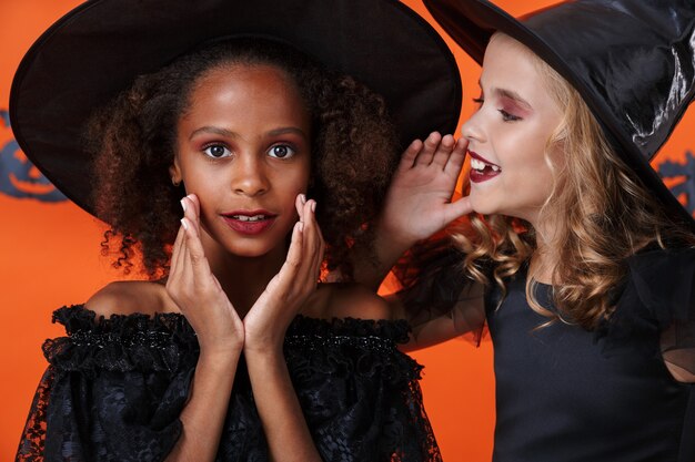 schattig meisje in zwart halloween-kostuum fluistert geheim tegen vriend in haar oor geïsoleerd over oranje pompoenmuur