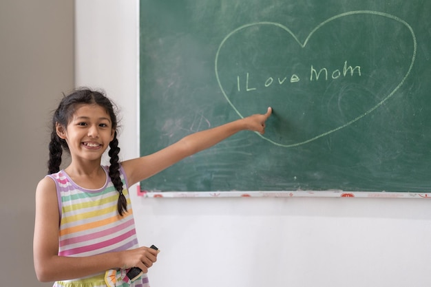 Schattig meisje met behulp van krijt tekening hart en liefde moeder schrijven op schoolbord muur in de klas op school