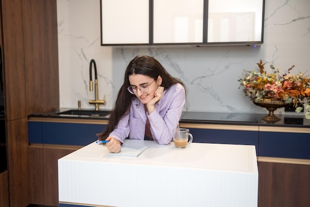 Schattig meisje met bril in de keuken koffie drinken en aantekeningen maken in een notitieboekje