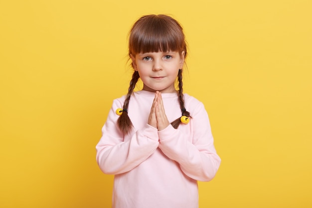 Schattig meisje op haar handpalmen te drukken, kind maken wens, poseren geïsoleerd op gele achtergrond, vrouwelijk kind uiting van verlangen, geloof, God vragen om te helpen, oprechte wens, klein meisje met staartjes bidden.