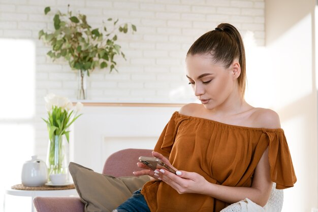 Schattig meisje van blanke etniciteit in casual kleding zit in een licht interieur in een stoel met een telefoon ...