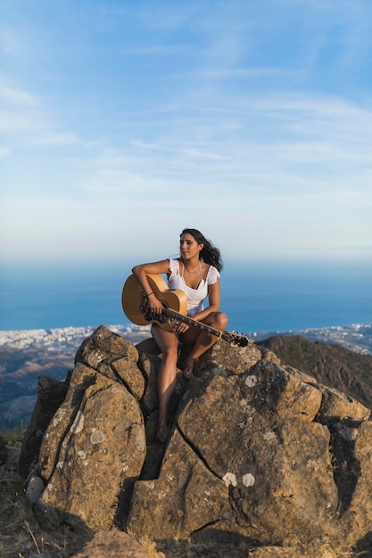 Schattig meisje zittend op de top van een berg met een gitaar in haar handen en zingend en glimlachend