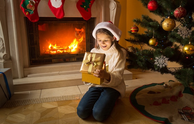 Schattig meisje zittend op de vloer bij open haard en kerstcadeau ontvangen in gouden doos