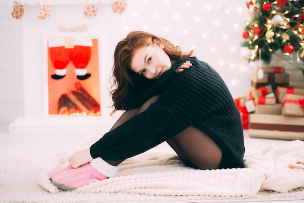 Schattig meisje zittend op het tapijt tegen de kerstboom en open haard in een gezellige trui