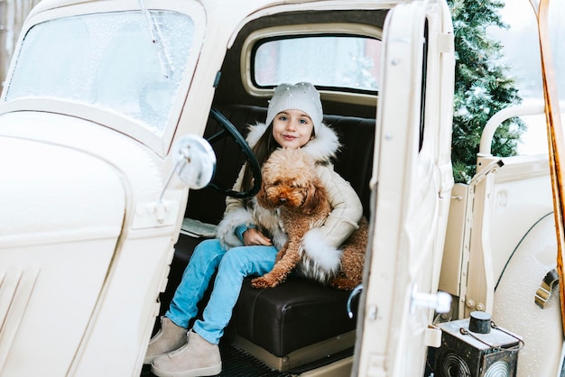 Schattig mooi Armeens meisje in winterjas en gebreide muts en huisdier poedel hond zitten in de buurt van beige retro pick-up truck ingericht voor Kerstmis en Nieuwjaar vintage interieur koffers ski's slee