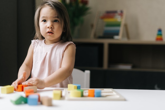 Schattig mooi meisje dat een witte jurk draagt, speelt met kleurrijk houten blokspeelgoed aan tafel in een gezellige kinderkamer