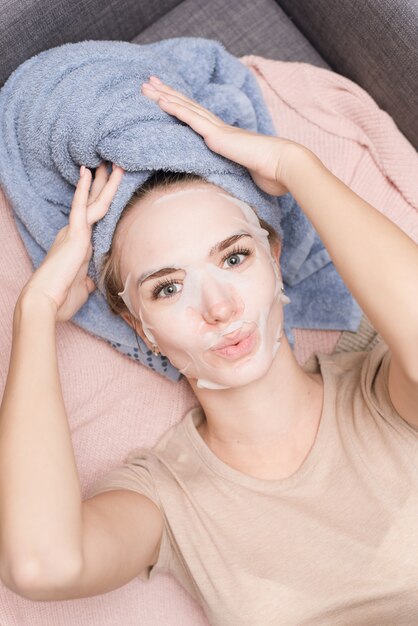 Schattig mooi meisje ligt op de bank in een wegwerp stof hydraterende masker en handdoek op haar hoofd en glimlachen. Thuiszorg, huidverzorging en anti-aging behandelingen