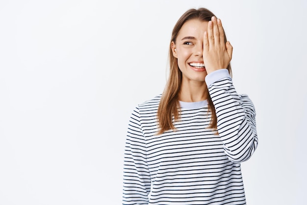 Schattig openhartig meisje met natuurlijk licht make-up bedekken de helft van het gezicht met palm en kijken naar links naar grafiek of logo staren advertentie staande op witte achtergrond