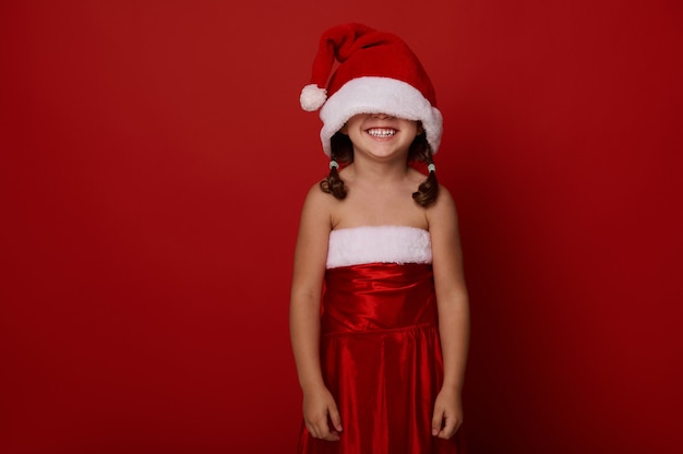 Schattig prachtig 4 jaar oud meisje, schattig kind in kerstman kleding en hoed die haar ogen bedekt, glimlacht brede glimlach poseren tegen rode achtergrond met kopie ruimte voor Kerstmis en Nieuwjaar advertentie