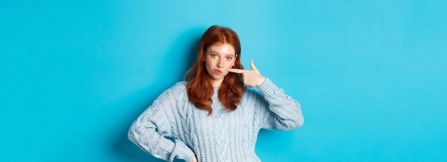 Foto schattig roodharig meisje in trui die haar wang porren starend naar camera brutaal staande over blauwe achtergrond