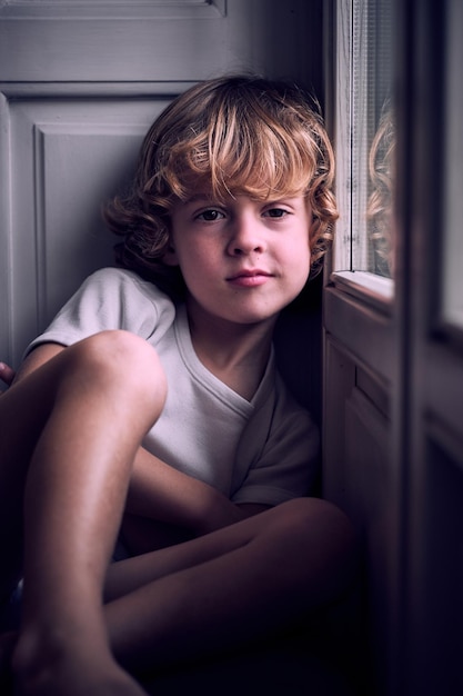 Schattig rustig jongetje met krullend blond haar in wit t-shirt leunend op het raam en kijkend naar de camera thuis