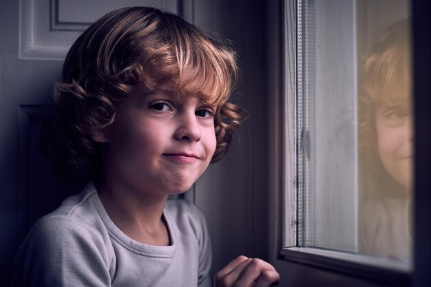 Schattig tevreden kind met krullend blond haar in t-shirt glimlachend en kijkend naar de camera terwijl hij overdag thuis bij het raam zit