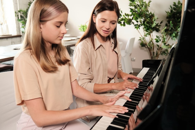 Schattig tienermeisje met lang blond haar, zittend bij piano naast haar moeder terwijl ze allebei muzikale dingen spelen in de woonkamer