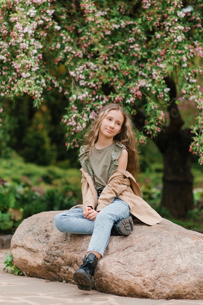 Schattig tienermeisje met lang bruin haar in een trenchcoat zit op een rots in het park en droomt