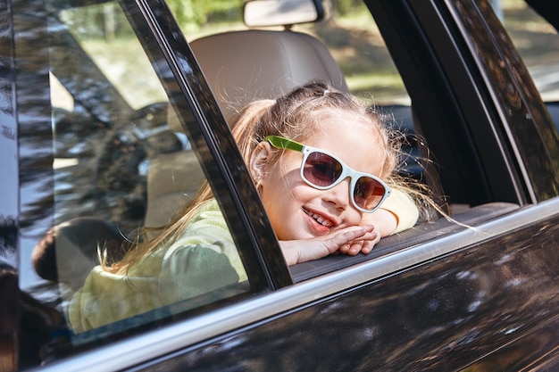 Schattig vrolijk klein kaukasisch meisje dat in de auto zit en uit het raam kijkt en glimlacht