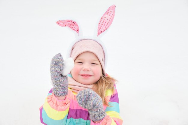 Schattig vrolijk kleutermeisje met caranale konijnenoren op haar hoofd speelt met een hart gemaakt van sneeuwsneeuw in april