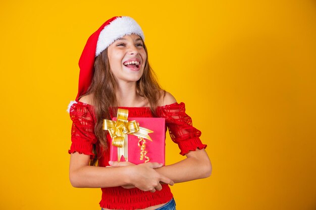 Schattig vrolijk meisje in een kerstmuts op een gekleurde achtergrond met een geschenk