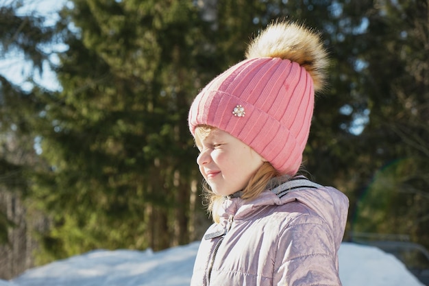 Schattig vrolijk peutermeisje in een besneeuwd bos, heldere lentezon, sneeuw in de lente, een meisje met een roze hoed loenst