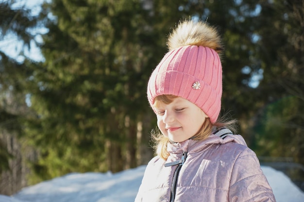 Schattig vrolijk peutermeisje in een besneeuwd bos, heldere lentezon, sneeuw in de lente, een meisje met een roze hoed loenst