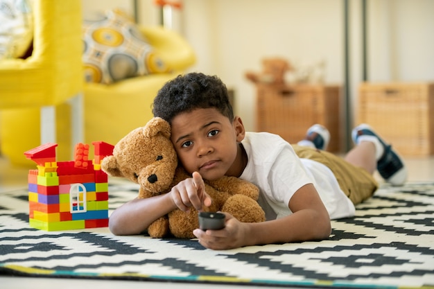 Schattige afrikaanse kleuter met afstandsbediening en bruine teddybeer wijzend op je liggend op de vloer van de woonkamer