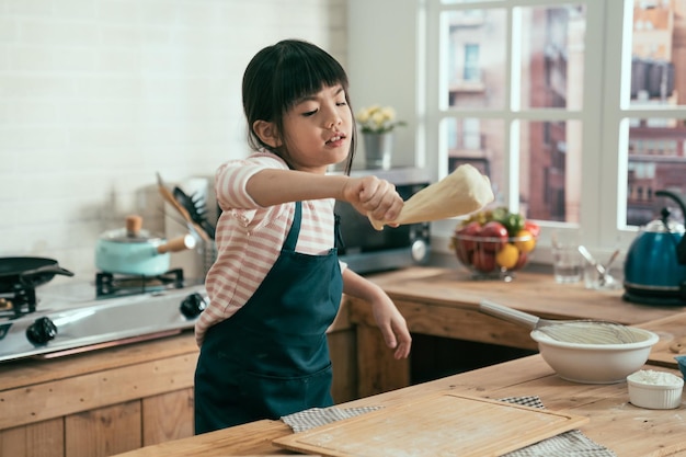 schattige aziatische chinese dochterchef-kok in schort die plezier heeft met het spelen van schommel met deeg voor het bakken van cake op moederdag in houten keuken. klein meisje dat handgemaakt brood maakt voor de paasvakantie geniet van diy.