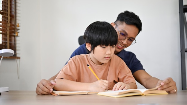 Schattige Aziatische kleine jongen die zich concentreert op zijn huiswerk terwijl zijn vader lesgeeft en helpt