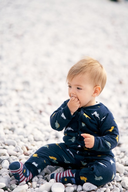 Schattige baby in blauwe overall zit op een kiezelstrand en heeft een kiezelsteen in zijn hand