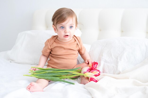 schattige baby in bruin katoenen rompertje zittend op bed met roze tulpen kind viert lentevakantie