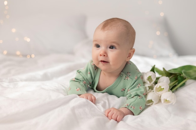 Schattige baby in een groene katoenen bodysuit thuis op een wit bed met tulpen. Lente, Moederdag, maart